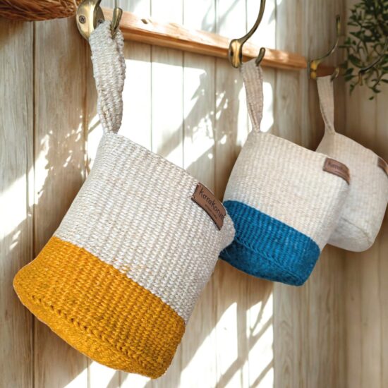 Handwoven sisal hanging baskets with twist handles — neutral with mustard base, neutral with indigo base, and natural — hanging on brass hooks in soft daylight.”