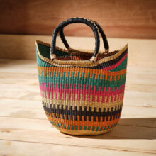 Ethically made woven grass market basket featuring teal, pink, orange and black ladder-stripe bands with leather-wrapped handles.