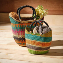 2 colourful handwoven market basket bags with leather wrapped handles filled with blankets and flowers on a wooden floor.