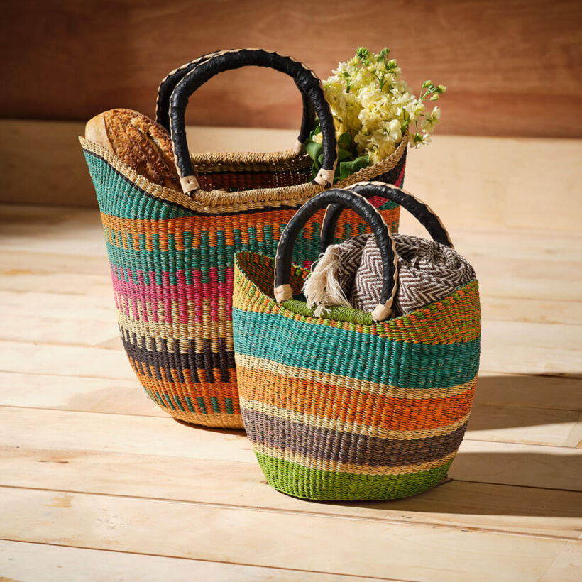 2 colourful handwoven market basket bags with leather wrapped handles filled with blankets and flowers on a wooden floor.