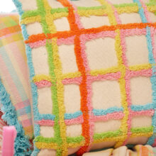 Close up of Handmade tufted check cushion cover in multicolour on a natural recycled cotton base, with tufted grid pattern in orange, pink, yellow, green and blue, finished with a blue tufted border. 45x45cm.