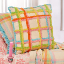 Handmade tufted check cushion cover in multicolour on a natural recycled cotton base, with tufted grid pattern in orange, pink, yellow, green and blue, finished with a blue tufted border. 45x45cm.