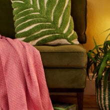 Hand tufted palm leaf cushion cover in green on a natural recycled cotton base, with a large frond design in tufted pile on a sofa with soft pink throw. 45x45cm, handmade in India.