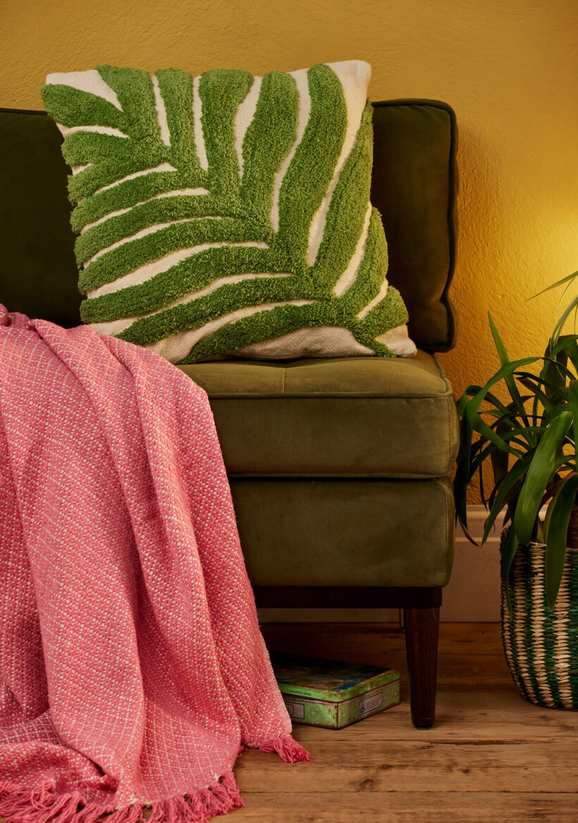 Hand tufted palm leaf cushion cover in green on a natural recycled cotton base, with a large frond design in tufted pile on a sofa with soft pink throw. 45x45cm, handmade in India.